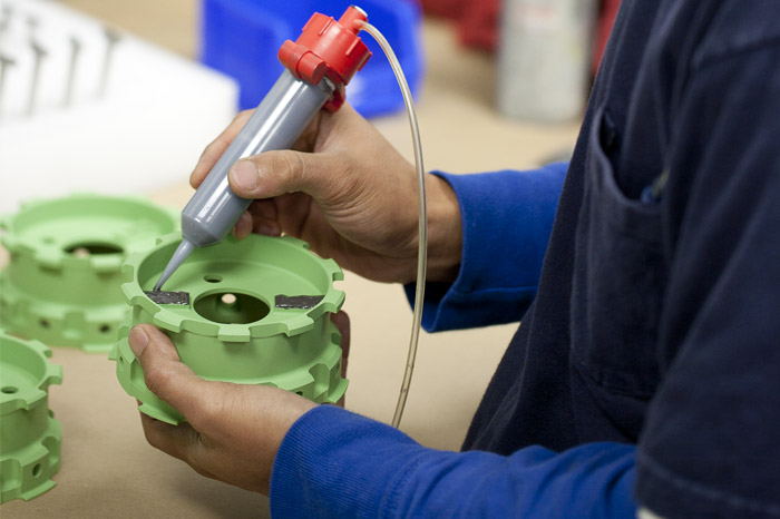 a person in the process of assembling or inspecting a green plastic component with the help of a gray and red handheld tool, which appears to be dispensing or applying a substance or performing a measurement. The setting suggests an assembly or quality control area within a manufacturing facility. The person's focus is on the task at hand, ensuring the correct application or measurement of the component, which may be part of a larger assembly in an industrial product.