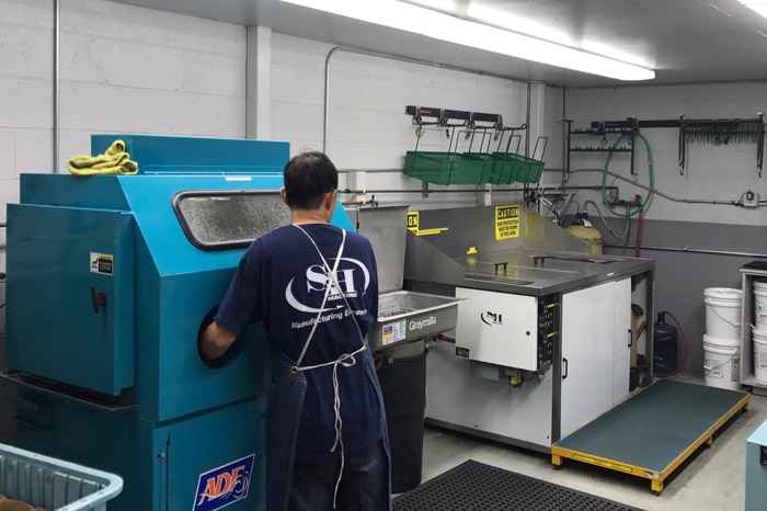 The image depicts an industrial setting where a person, wearing a blue t-shirt with a logo on the back, is operating machinery used for cleaning or finishing parts. On the left, there's a large blue machine, possibly a parts washer or deburring machine, branded with 