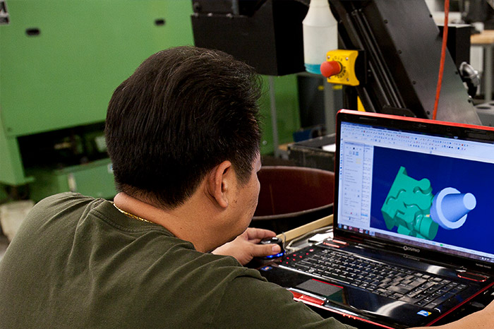 a person working on a laptop with a 3D CAD (Computer-Aided Design) model on the screen, suggesting that they are engaged in an engineering design or review process. The CAD model appears to be a part with a complex shape, likely for manufacturing. In the background, there is industrial machinery, possibly a CNC machine, indicating that this setting is a manufacturing facility or machine shop. The person appears focused on their work, possibly making adjustments to the design or preparing the model for machining. This scene represents the integration of modern design technology in a manufacturing environment.