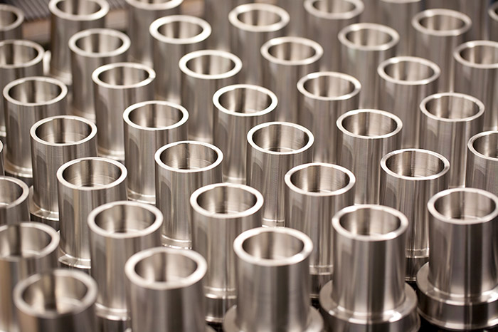 a collection of cylindrical metal components closely arranged together. Each piece has a hollow center, suggesting that they may be some form of sleeves, spacers, or bushings. The parts have a reflective metallic finish, which could indicate that they are made from stainless steel or aluminum. Their uniformity and precision suggest they were produced through a process like CNC turning or milling. This array of components could be part of a stock inventory in a machine shop, awaiting further processing or assembly. The repetition of shapes and the consistency in manufacturing reflect a high level of quality control.