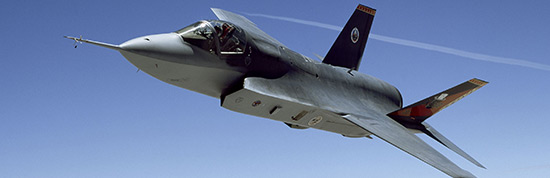 a fighter jet in flight against a clear blue sky. The jet has a single tail fin and distinctive air intakes on the sides, indicating it is a military aircraft designed for speed and maneuverability. Its sleek aerodynamic shape is typical of modern combat aircraft, and there is a visible cockpit where the pilot operates the jet. The presence of markings and insignia on the tail would usually identify the aircraft's national origin or the military branch it belongs to.