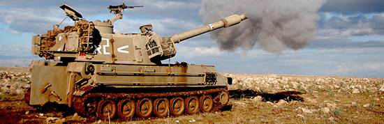 a military tank in a firing position in a rocky, barren landscape. The tank is tracked, armored, and equipped with a large caliber cannon, which is in the process of firing, as evidenced by the smoke and the visible blast from the barrel. The design of the tank, with its robust tracks and heavily armored turret, suggests it is built for direct combat roles, providing firepower and survivability on the battlefield. The setting appears to be a rough, outdoor terrain, likely simulating a combat environment.