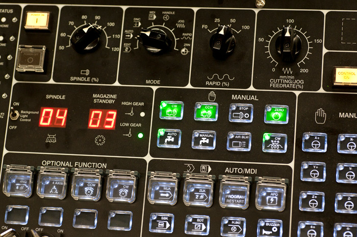 a control panel, likely from a CNC (Computer Numerical Control) machine given the specific controls and digital readouts. It has various buttons and switches for different functions such as spindle speed control, mode selection (auto, manual, MDI), feed rate override, and rapid movement percentage. The panel also includes optional functions for axis control, an emergency stop button, and settings for the spindle gear (high/low). LED indicators provide visual feedback for the operator on the status of different machine functions. The numerical displays are currently showing 