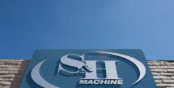 a bright, clear day with a blue sky and few clouds. The perspective is looking up toward the top of a building, where a large sign is mounted on a textured stone facade. The sign features the logo and name of "S&H Machine," suggesting that this is a manufacturing or machining company. The logo includes a stylized letter 'S' that curves around a central point, creating a dynamic and modern look. The angle of the photo gives a sense of the company's pride in its American identity and its place within the manufacturing industry.