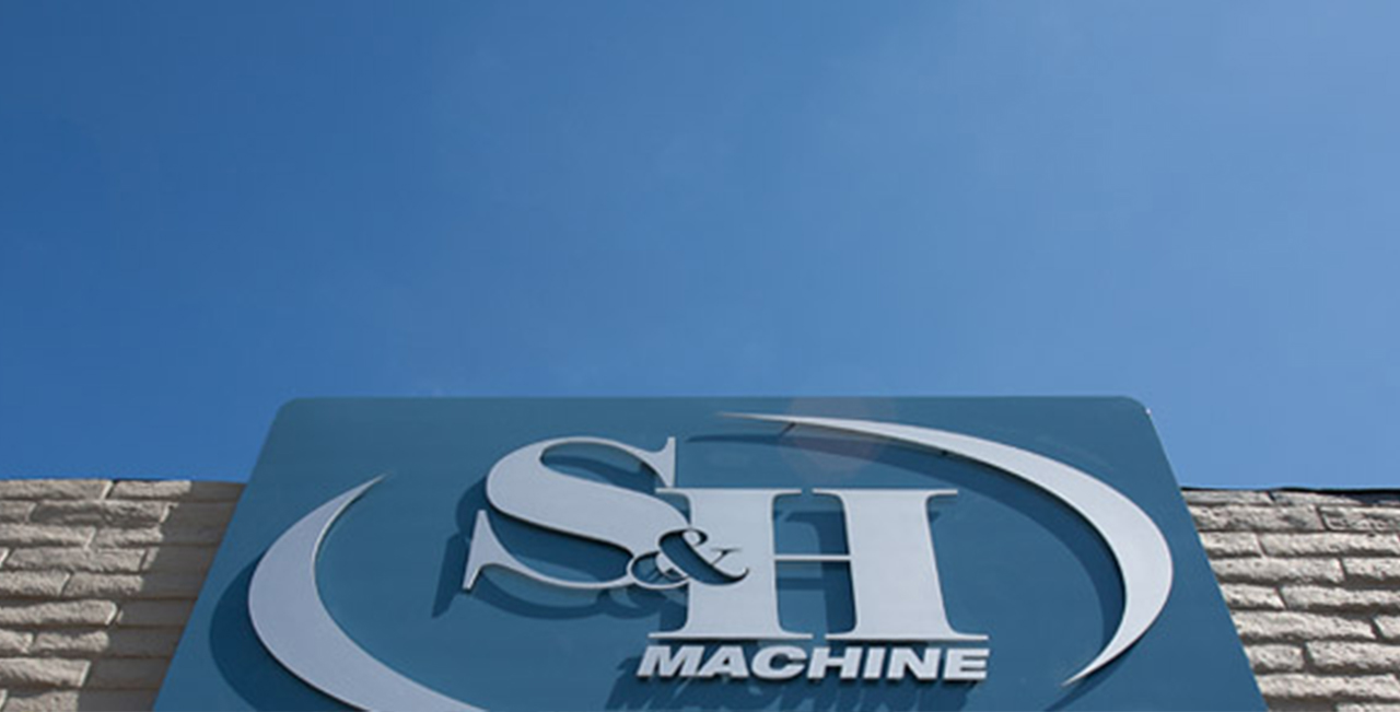 a bright, clear day with a blue sky and few clouds. The perspective is looking up toward the top of a building, where a large sign is mounted on a textured stone facade. The sign features the logo and name of 