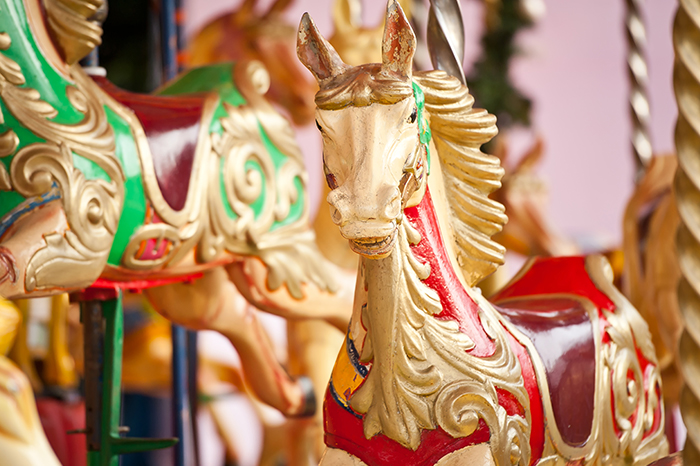 a carousel horse, which is part of a merry-go-round, a ride commonly found at amusement parks and fairs. The horse is ornately decorated with traditional carousel artistry, featuring gold, red, and green colors with intricate designs and flourishes that suggest an older, classic style. The pole in the center suggests it is one of the ride's moving figures, designed to go up and down as the carousel turns. The blurred background with another similar figure hints at the larger context of the carousel. This image evokes nostalgia and the simple joy of childhood amusement park experiences.