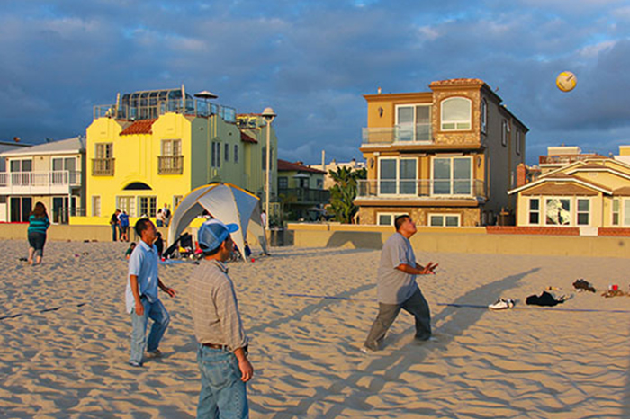 a beach scene during what appears to be late afternoon, given the warm, golden lighting. Several individuals are casually playing beach volleyball, with one person in the foreground hitting the ball. Onlookers and other beachgoers are present, some watching the game, others engaged in their own activities. In the background, there are colorful beachfront houses, which add a vibrant backdrop to the leisurely setting. The overall atmosphere of the picture is one of relaxation and enjoyment of a beautiful day on the sand, typical of a seaside community lifestyle or a vacation setting.