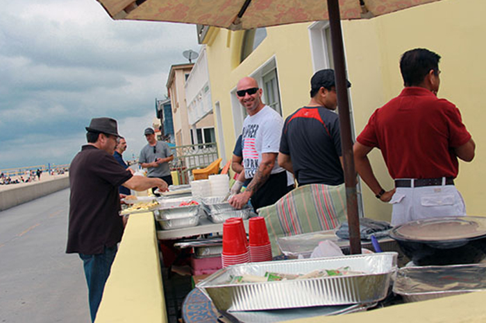 Several people are standing around a serving table that is covered with aluminum trays of food, stacks of plates, and red plastic cups, suggesting a buffet-style service. A man in the center is smiling and engaging with someone, indicating a relaxed, friendly atmosphere. The presence of umbrellas hints that it might be a sunny day or provides protection in case of light rain. In the background, there is a view of an open area that could be a beach or a wide promenade, adding to the leisurely ambiance. This scene likely represents a company or community event meant to provide a break from work, focusing on socialization and enjoyment.