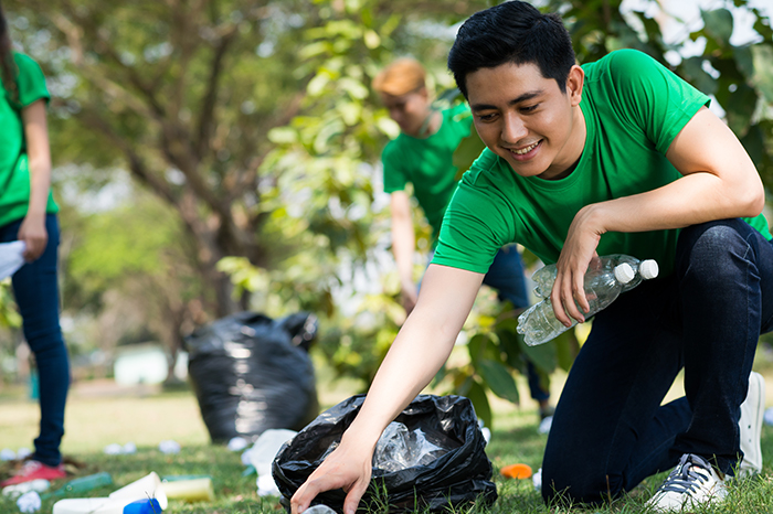 a group of individuals participating in an outdoor cleanup activity. They are wearing matching green t-shirts, which often signifies team or organizational unity, suggesting this could be a company-sponsored volunteer event. The focus is on a young man squatting down to pick up a plastic bottle, placing it into a black trash bag. His smile indicates a positive and enthusiastic approach to the task. In the background, others are also collecting trash, and there appear to be several filled garbage bags already. This scene conveys a sense of community service and environmental responsibility, highlighting an initiative to keep public spaces clean and promote ecological awareness.