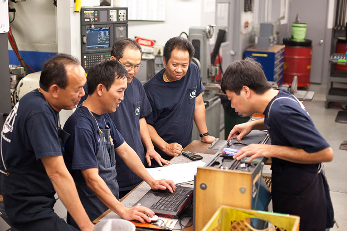 a group of five men focused on a task in what appears to be a workshop or manufacturing environment. They are gathered around a workbench where a laptop and some papers are visible, suggesting they may be discussing plans or data related to machining. The presence of a CNC machine in the background indicates that this could be a hands-on training session or a problem-solving discussion. All individuals are dressed in dark blue work uniforms with the "S&H MACHINE" logo, which implies they are part of the same team or company, engaged in skilled manufacturing work, possibly as part of an apprenticeship or continuous professional development program.