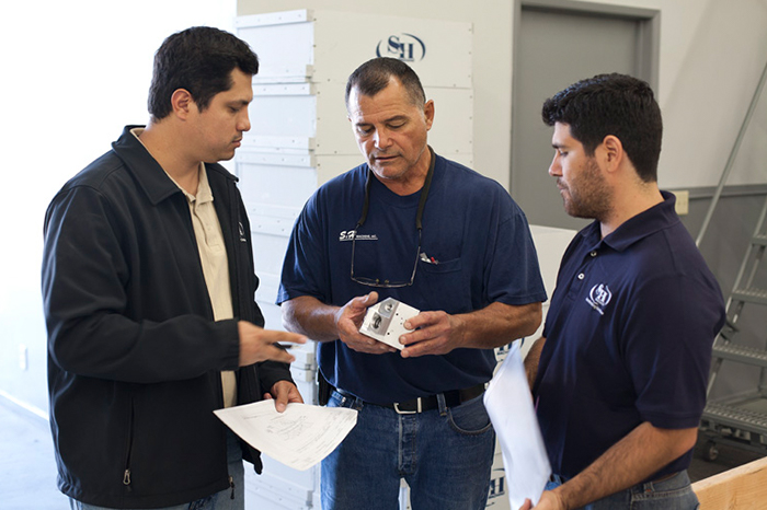 Three men looking at a precision machined part held by the man in the center. The other two men hold plans or documents while the center man explains a feature of the part suggesting training.