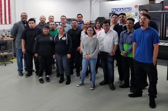 A group of students from LAVCC standing in a group while visiting the S&H Machine facility. They are all wearing safety glasses. A CNC machine and an American flag can be seen in the background.