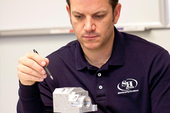 a focused man wearing a dark polo shirt embroidered with the logo "S&H MACHINE Manufacturing Excellence". He is closely examining a metallic mechanical component, possibly during an inspection or quality control process. The man holds a pen in his hand, suggesting he may be about to take notes or mark the part. The setting looks like a workshop or educational environment due to the plain background and fluorescent lighting, which is common in manufacturing facilities and training areas. This could be indicative of hands-on instruction or mentorship in precision manufacturing or engineering.