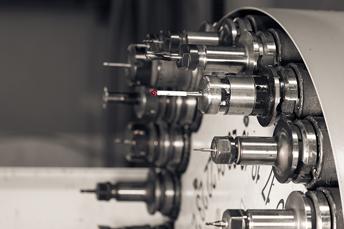 a tool turret from a CNC (Computer Numerical Control) lathe or milling machine. The turret holds various cutting tools and can rotate to bring each tool into the working position as needed during machining operations. The focused laser point might be part of a tool pre-setter system used to measure the tool's geometry. This is typically used in precision manufacturing settings where accurate machining is critical. The photo has a narrow depth of field, with the first few tools in focus, gradually blurring further down the line, highlighting the precision required in such environments. The color tone suggests it's been taken with a filter or edited for a more dramatic, professional look, emphasizing the intricacy of the machinery.