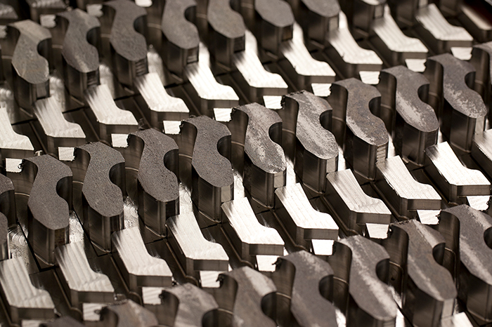 a large number of identical metal components that are likely manufactured using a precision machining process. They are organized in rows and columns, with a uniform appearance, suggesting they are part of a batch production. The parts have a complex geometry with curved and flat surfaces, and the shine on the edges indicates a finished machining process. This image could be used in industrial contexts to showcase manufacturing capabilities, precision, and uniformity in production. It gives a sense of scale of manufacturing operations where numerous parts are produced to exact specifications, which could be essential for industries that require high-quality machine components, such as aerospace or automotive sectors.