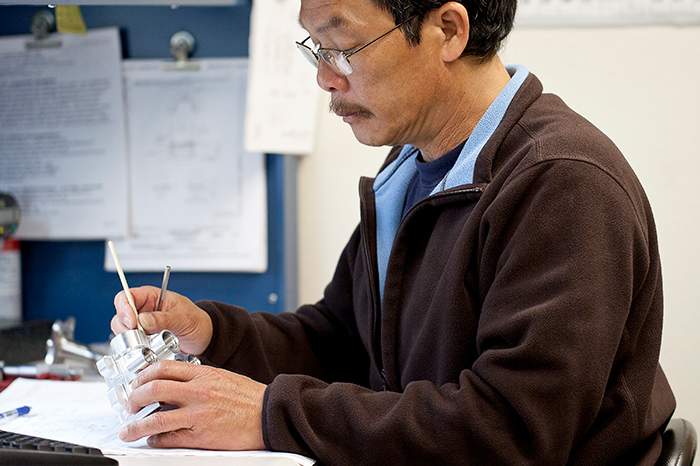 an individual concentrating on inspecting or assembling a complex mechanical part, likely in a quality control or engineering context. The person is sitting at a desk with papers that may include technical drawings or inspection reports. They are using a precision tool, possibly a caliper or a similar measuring device, indicating a meticulous approach to ensuring the part meets strict specifications. The setting is an office-like environment within an industrial or manufacturing facility, emphasizing the detailed and focused nature of the task at hand.